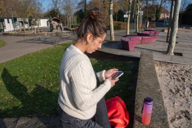 Dışarıda oturan bir kadın, güneşli bir günde halka açık bir parkta dinlenirken iletişim ve bilgi için kablosuz teknolojiyi kullanarak akıllı telefon ekranına odaklanıyor.