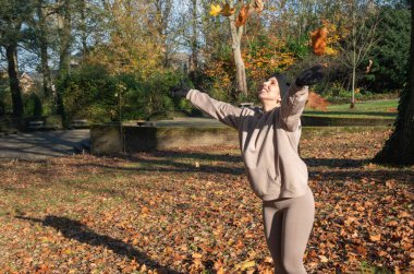Gülümseyen ve renkli sonbahar yapraklarını havaya fırlatan genç bir kadın, düşen yeşillikler ve sıcak güneş ışığının ortasındaki halka açık bir parkta neşe ve özgürlük yaşıyor.