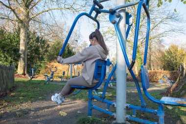 Sunny Park 'ta açık hava göğüs pres makinesi kullanan bir kadın yeşil çimenler ve ağaçların arasında, yaz sporu, yan ve arka görüş alanlarında dayanıklılık eğitimi ve aktif sağlıklı yaşam tarzına odaklanmış.