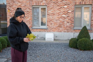 Sıcak kapüşonlu ve gözlüklü bir kadın renkli akçaağaç yaprakları tutuyor, sessiz bir banliyö bahçesinde tuğla bir duvarın yanında duruyor, sakince sonbahar yapraklarının ve mevsimsel değişimin tadını çıkarıyor.