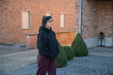 Spor kıyafetleri içinde, siyah bir kapüşonlu, bordo eşofman altı, gözlük ve saç bandı takan bir kadın, rahat ve günlük yaşam tarzını yansıtıyor.