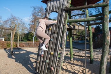 Genç bir kadın, kum kaplı bir yüzey üzerindeki idman sırasında tutunma ve hareket odaklı kaya duvarına benzeyen ahşap bir oyun alanına aktif olarak tırmanıyor.