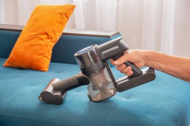 Güçlü bir kablosuz el süpürgesi tutan insanlar, evdeki oturma odasındaki turuncu yastıklı çağdaş mavi kumaş kanepesinden toz ve döküntüleri etkin bir şekilde kaldırıyorlar.