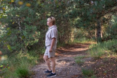 Olgun bir kadın, sonsuz yeşil bir ormanda huzurlu bir toprak yolda duruyor, günlük spor giysileri giyiyor, doğayı yansıtıyor ve sessiz açık havanın tadını çıkarıyor.