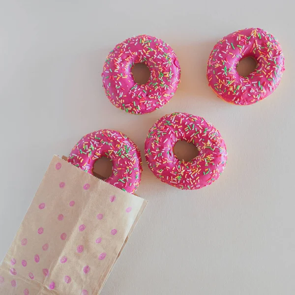 Four bright pink glazed doughnuts with sprinkles and a craft bag on a light background