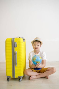 Çocuk elinde küreyle sarı bir bavulun yanında yerde oturuyor..