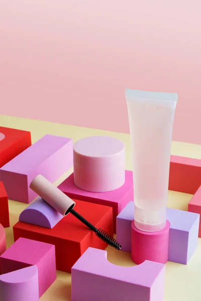 Decorative cosmetics kit. Jar and bottle of cosmetic cream,eyelash brush (mascara). Cosmetic products on colourful cubes on yellow and pink background. Beauty and morning skin care concept.