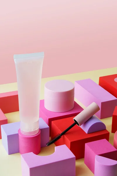 Decorative cosmetics kit. Jar and bottle of cosmetic cream,eyelash brush (mascara). Cosmetic products on colourful cubes on yellow and pink background. Beauty and morning skin care concept.