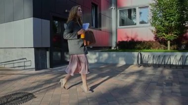 İş kadını, modern ofis binalarının yakınında elinde kahve ve çanta tutarak yürüyor..