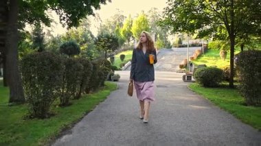 İş kadını, elinde kahve fincanı ve çantasıyla şehir parkında kasten yürüyor..