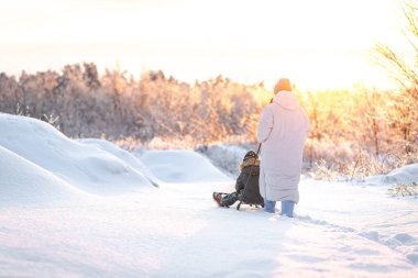 Genç bir anne, oğlunu gün batımına karşı karlı bir alanda kızakla kaymaya götürür. Kış, annelik ve kış aktiviteleri kavramı..