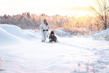 Genç bir anne, oğlunu gün batımına karşı karlı bir alanda kızakla kaymaya götürür. Kış, annelik ve kış aktiviteleri kavramı..