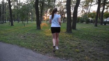 Gün batımında şehir parkında kameradan kaçan genç bir kadın. Ilık akşam ışığı ile yavaş çekim geri bakış sinematik ve ilham verici bir atmosfer yaratır. Fitness, özgürlük ve sağlıklı yaşam tarzı kavramları için ideal..