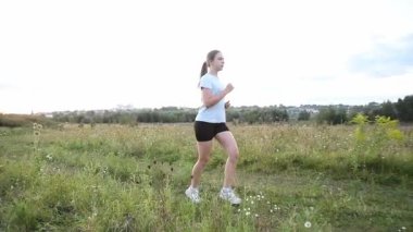 Gün batımında, dikey kamera hareketiyle kırsal toprak yolda koşan genç bir kadın. Kamera uzaklaşırken yavaş çekim kenarı görüntüsü dinamik sinematik perspektif yaratır. Fitness, dayanıklılık ve özgürlük kavramları için idealdir..