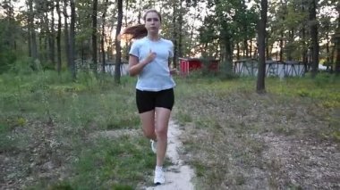 Gün batımında hareket eden bir kamera görüntüsüyle park yolunda koşan genç bir kadın. Öndeki izleme hareketi yavaş yavaş bir taraftan diğerine kayarak dinamik sinematik perspektif yaratıyor. Fitness, motivasyon ve aktif yaşam tarzı kavramları için ideal.