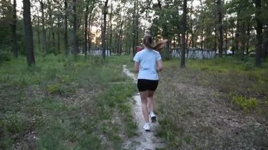 Park yolunda yavaş çekimde gün batımına doğru koşan genç bir kadın. Ilık altın ışıkla çekilen arka plan takibi ilham verici ve sinematik bir atmosfer yaratır. Motivasyon, özgürlük ve sağlıklı yaşam tarzı kavramları için ideal..