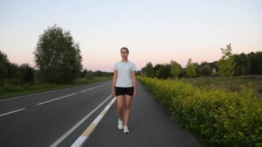 Gün batımında yol kenarındaki kaldırımda kablosuz kulaklık takan genç bir kadın. Yavaş çekim alçak açı çekim özgüven ve modern şehir yaşam tarzı atmosferi yaratır. Sağlık, rutin ve aktif şehir kavramları için ideal..