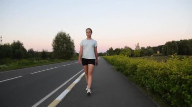 Genç bir kadın gün batımında kaldırımda yürüyor ve kablosuz kulaklık takarak koşmaya başlıyor. Yavaş çekim düşük açı çekimi güçlü ve motive edici kentsel zindelik atmosferi yaratır. Kararlılık, ilerleme ve aktif yaşam tarzı için ideal.