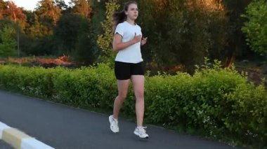 Gün batımında yol kenarında, kablosuz kulaklık takan genç bir kadın. Sıcak akşam ışığı ve park arka planıyla gerçek zamanlı çekim yapmak dinamik kentsel fitness atmosferi yaratır. Aktif yaşam tarzı ve teknoloji kavramları için ideal.