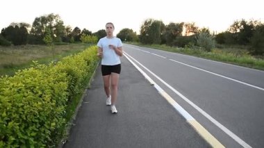 Su içerken ve günbatımında kablosuz kulaklık takarken kaldırım boyunca kameraya doğru yürüyen genç bir kadın. Doğal hareketle dinamik ön izleme çekimi modern kentsel zindelik atmosferi yaratır. Sağlık, iyileşme ve aktif yaşam tarzı için ideal..