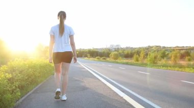 Genç bir kadın sıcak gün batımı ışığında kaldırımda yürüyor. Yavaş çekim geri izleme çekimi yumuşak sinematik şehir yaşam tarzı atmosferi yaratır. Sağlık, rutin ve ilham verici günlük kavramlar için idealdir..