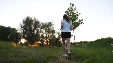 Genç bir kadın çamur parkında gün batımına doğru koşuyor. Çim seviyesinin yakınından çekilmiş. Yavaş çekim geri izleme çekimi güçlü bir sinematik derinlik ve ilham verici bir atmosfer yaratır. Motivasyon, dayanıklılık, kişisel büyüme kavramları için ideal.