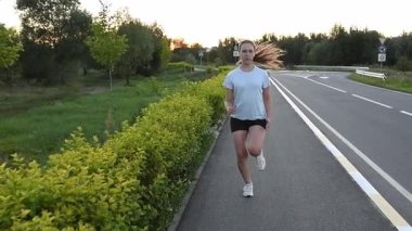 Gün batımında kaldırımda kameraya doğru koşan genç bir kadın kablosuz kulaklık takıyor. Yavaş çekim hızı, gücü ve dinamik kentsel zindelik atmosferini vurgular. Performans, dayanıklılık ve aktif yaşam tarzı kavramları için ideal.