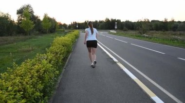Gün batımında, kablosuz kulaklık takıp kaldırımda kameradan kaçan genç bir kadın. Yavaş çekim, uzun güçlü adımları ve dinamik kentsel zindelik atmosferini vurgular. Dayanıklılık, güç ve performans kavramları için idealdir..