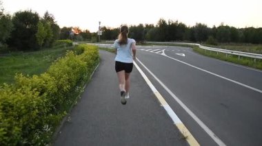 Gün batımında, kablosuz kulaklık takıp kaldırımda kameradan kaçan genç bir kadın. Yavaş çekim, uzun güçlü adımları ve dinamik kentsel zindelik atmosferini vurgular. Dayanıklılık, güç ve performans kavramları için idealdir..