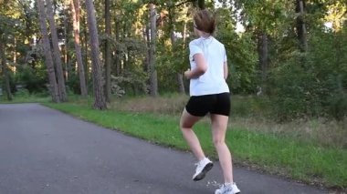 Genç bir kadın kaldırımlı park yolunda kameradan kaçıyor. Ağaçlar arasında ağır çekimde. İzleme çekimi doğal derinlik ve dinamik açık hava fitness atmosferi yaratır. Dayanıklılık, sağlık ve aktif yaşam tarzı kavramları için idealdir..