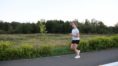 Park arka planında gerçek zamanlı olarak kaldırım kenarında koşan genç bir kadın. İzleme çekimi yavaş yavaş önden yana ve arkaya doğru kayar, dinamik kentsel zindelik atmosferi yaratır.