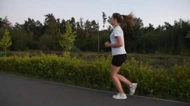 Park yolunda yavaş çekimde koşan genç bir kadın kameranın bir yandan diğerine geçişi gibi. İzleme hareketi dinamik derinlik ve açık hava fitness atmosferi yaratır. Dayanıklılık, yolculuk ve aktif yaşam tarzı kavramları için ideal.