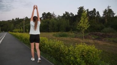 Gün batımında kameradan uzaklaşan genç bir kadın derin derin nefes alıyor ve kollarını kaldırıyor ve indiriyor. Yavaş çekim geri bakış sakin ve bilinçli kentsel refah ortamı yaratır. İyileşme, rahatlama ve sağlıklı yaşam tarzı için ideal..