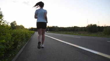 Gün batımında kaldırımda kameradan kaçan genç bir kadın düşük yerden yavaş çekimde filme alındı. Geri izleme çekimi güçlü adımları ve dinamik kentsel zindelik atmosferini vurgular. Dayanıklılık, güç ve aktif yaşam tarzı kavramları için ideal..