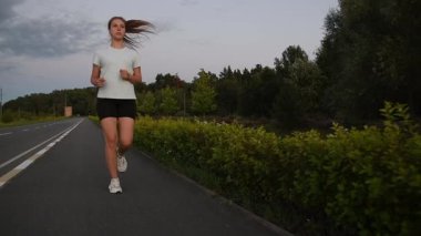 Gün batımında kaldırımda kameraya doğru koşan genç bir kadın alçaktan çekilmiş. Ön izleme çekimi güçlü hareketi ve dinamik kentsel zindelik atmosferini vurguluyor. Performans, dayanıklılık ve motivasyon kavramları için ideal.