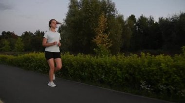 Gün batımında kaldırımda ağır çekimde koşan genç bir kadın çapraz takip hareketiyle. Kamera yavaş yavaş bir açıyla uzaklaşıyor, dinamik derinlik ve kentsel zindelik atmosferi yaratıyor. Dayanıklılık, performans, aktif yaşam tarzı için ideal