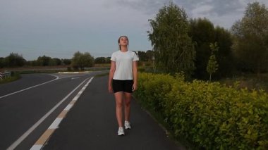 Gün batımında kaldırımda kameraya doğru yürüyen genç bir kadın derin bir nefes alıyor. Sıcak akşam ışığında yavaş çekim yapmak sakin ve düşünceli bir şehir atmosferi yaratır. İyileşme, sağIık ve sağlıklı yaşam tarzı kavramları için ideal..