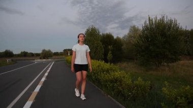 Gün batımında kaldırımda kameraya doğru yürüyen genç bir kadın derin bir nefes alıyor. Sıcak akşam ışığında yavaş çekim yapmak sakin ve düşünceli bir şehir atmosferi yaratır. İyileşme, sağIık ve sağlıklı yaşam tarzı kavramları için ideal..