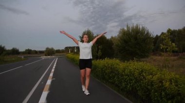 Genç bir kadın gün batımında kaldırımda kameraya doğru yürürken kollarını kaldırıyor ve indiriyor ve derin nefes alıyor. Yavaş çekim sakin ve dikkatli kentsel zindelik ortamı yaratır. İyileşme, nefes alma ve sağlıklı yaşam tarzı için ideal..