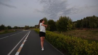 Gün batımında yol kenarında, omuzlarını açarken kameraya doğru yürüyen genç bir kadın. Sıcak akşam ışığında yavaş çekim sakin ve odaklanmış kentsel zindelik atmosferi yaratır. İyileşme, ısınma ve sağlıklı yaşam için ideal..