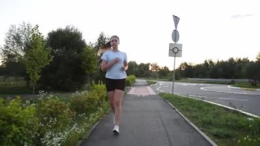 Gerçek zamanlı olarak kaldırım kenarında kameraya doğru koşan genç bir kadın. Ön izleme çekimi doğal hareketi ve enerjik kentsel zindelik atmosferini yakalar. Dayanıklılık, performans ve aktif yaşam tarzı kavramları için ideal.