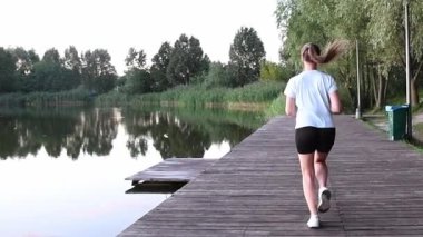 Ahşap göl kenarı rıhtımında ağır çekimde koşan genç bir kadın, arkadan çekilmiş. Arka plan görüntüsü, dinamik hareketi ve deniz manzaralı atmosferi yakalar. Özgürlük, dayanıklılık ve aktif yaşam tarzı kavramları için ideal..