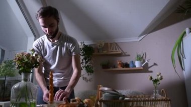 Mutfak masasında kahvaltıyı hazırlayan adam, kruvasan sandviçlere taze sebzeler eklemek için bir paket krem peynir açıyor. Ev yapımı yemek hazırlama ve şefkatli jestlere odaklanmış rahat bir sabah hayatı..