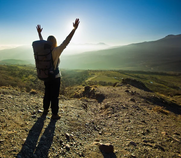 Menina-mochileiro com as mãos para cima nas montanhas contra o sol — Fotografia de Stock