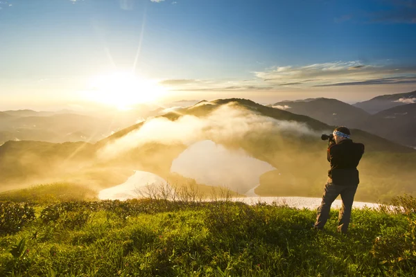 Homme sur une colline au coucher du soleil avec appareil photo faisant photo — Photo