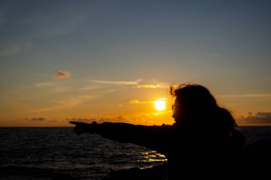 Güneş gözlüklü bir kadın batan güneşle denize uzak bir hedefi işaret ediyor. Fotoğraf batan güneşin atmosferik ışığı altında çekildi..