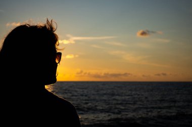 Güneş gözlüklü bir kadın yüzünü denizin üzerindeki batan güneşe çeviriyor. Fotoğraf batan güneşin atmosferik ışığı altında çekildi..