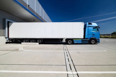 Lojistik merkezinin avlusuna park etmiş beyaz bir karavanı olan mavi bir kamyon. Kamyon yüklenmeyi ya da boşaltılmayı bekliyor. Fotoğraf güneşli bir günde çekildi..