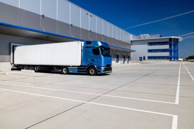 Lojistik merkezinin avlusuna park etmiş beyaz bir karavanı olan mavi bir kamyon. Kamyon yüklenmeyi ya da boşaltılmayı bekliyor. Fotoğraf güneşli bir günde çekildi..