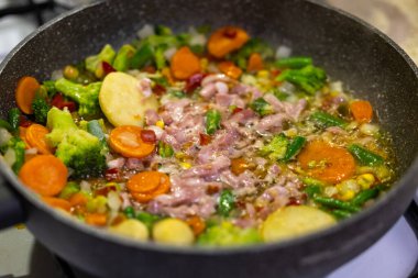 Renkli bir sebze ve pastırma karışımı bir tavada hazırlanıyor. Çanağın malzemelerinin statik yakın çekimi, yapay ışık altında çekiliyor..